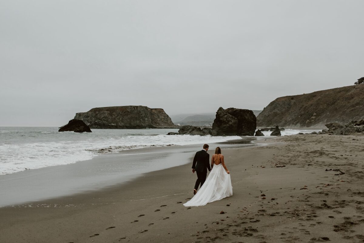 A couple walk along a beach on an overcast day. The groom is wearing a black suit and the bride is wearing a white lace dress with a long train. The beach is empty, with big rocks jutting out of the water. Discover how to choose a California elopement location that aligns with your love story, values, and sense of place in this post.