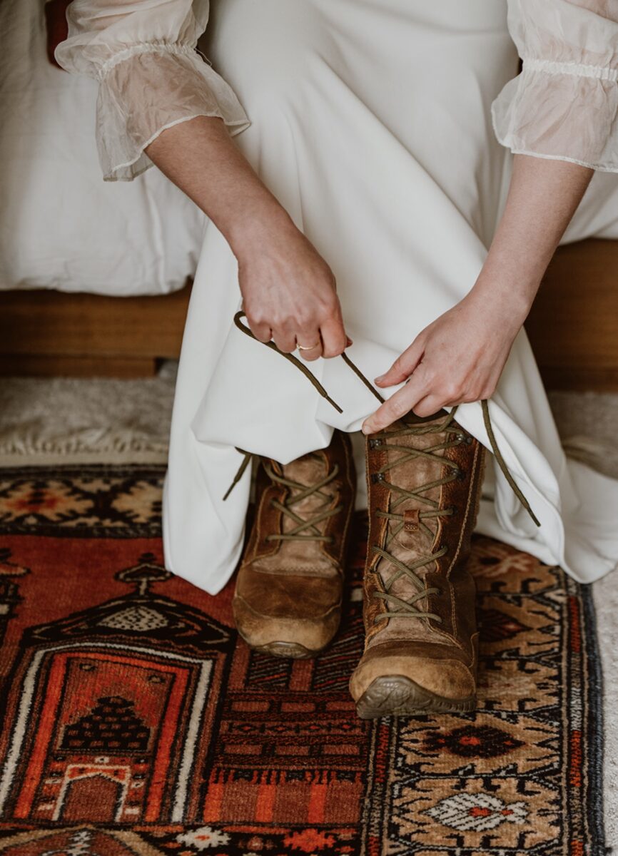A bride sits on a bed over a red Turkish rug and leans down to lace her hiking boots under her white floor-length dress. Check out this post for elopement outfit ideas for all bodies and styles—designed for beauty, comfort, and presence in any setting.