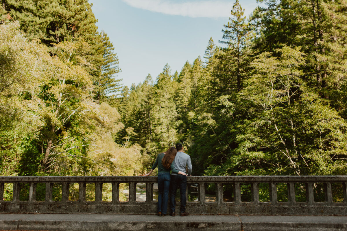 A couple stands together on a bridge lookout out over a creek in the forest; one partner has her head on the other's shoulder and a hand around his waist. Discover how to choose a California elopement location that aligns with your love story, values, and sense of place in this post.