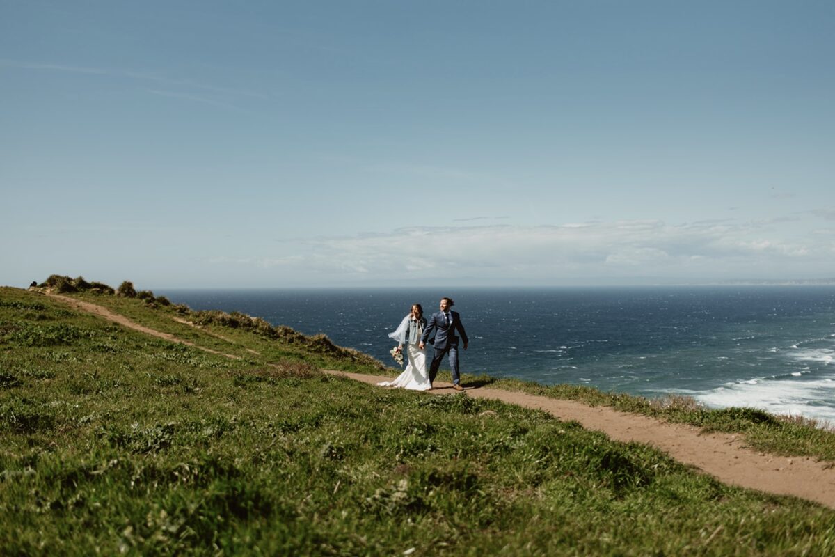 A couple walks in the sun on a bluff above the ocean. The path is bordered by green grass and the sky is clear. The groom wears a navy suit and the bride wears a white dress with a veil and jean jacket. Learn how to write wedding vows with prompts and emotional guidance to help you feel clear, calm, and connected—without sounding cliché.