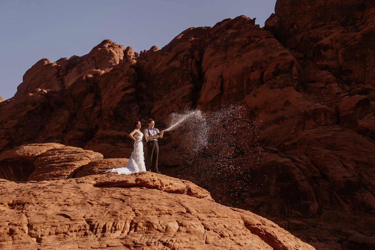 A couple pops a bottle of champagne in the desert, laughing as the spray is highlighted in the morning sun on the orange sandstone rock. Eloping doesn’t have to mean hiking or secrecy. Let’s debunk 10 myths and explore what your wedding day could actually feel like.