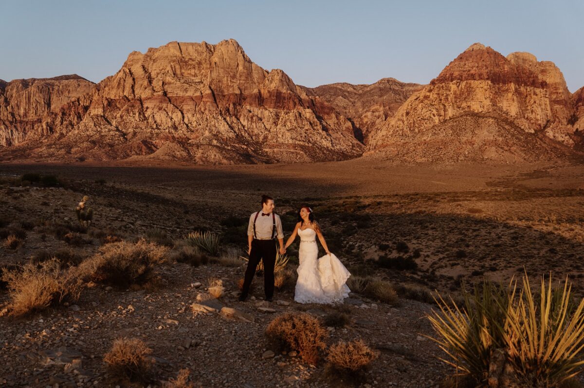 A couple walks hand in hand in the desert scrub at forest, laughing and looking at each other. The groom is wearing suspenders and a bowtie, and the bride is wearing a strapless white gown. There are towering rock formations behind them lit up in the red-orange light of sunrise. Discover how to choose a California elopement location that aligns with your love story, values, and sense of place in this post.