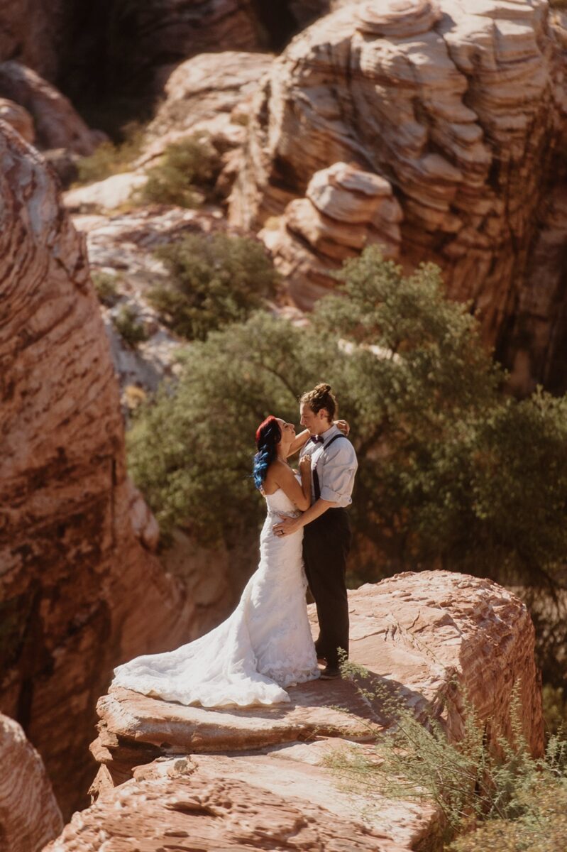 A couple in wedding clothes stand on an orange sandstone formation above a small canyon with trees in it. The groom wears suspenders and a bowtie, and the bride wears a strapless white gown with a train. Eloping doesn’t have to mean hiking or secrecy. Let’s debunk 10 myths and explore what your wedding day could actually feel like.