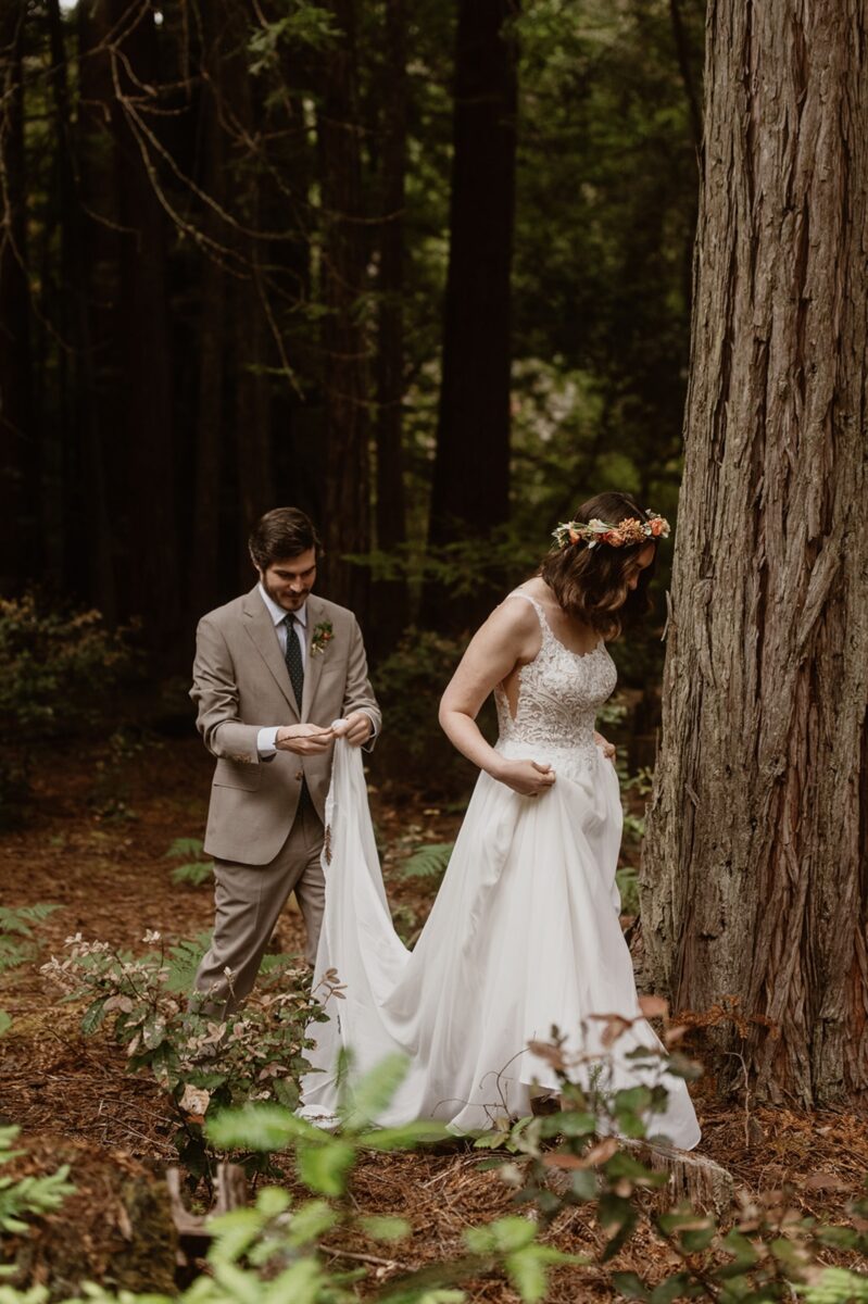 A couple walks through a redwood forest on their wedding day. The bride leads in a white dress and flower crown, while the groom follows in a beige suit picking sticks out of her train as they walk. In this post are gentle ways to tell family you're eloping—plus emotional tips, real scripts, and ways to honor your loved ones without losing yourself.