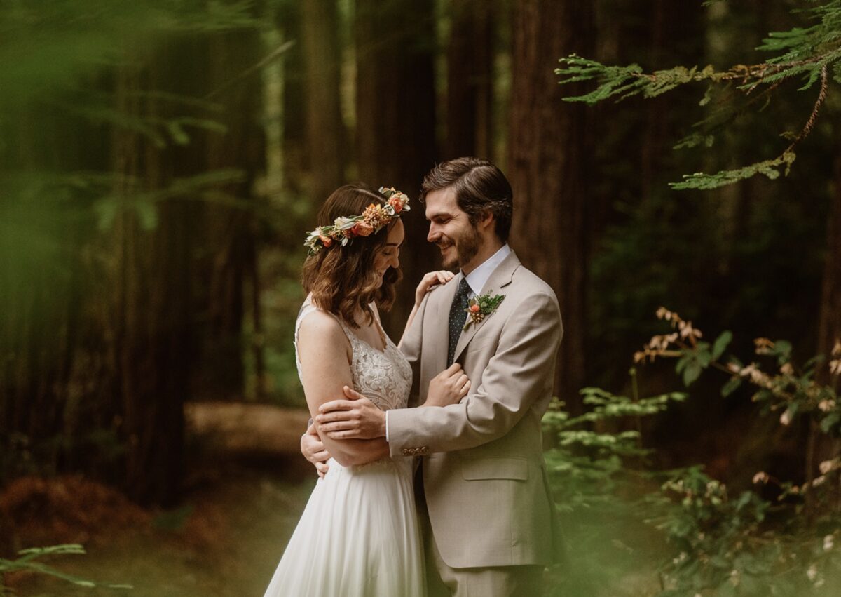 A couple in wedding clothes stands laughing with their heads close together in the redwood forest. The groom wears a beige suit, and the bride wears a lace-bodice wedding gown with an orange and yellow fresh flower crown. Check out this post for elopement outfit ideas for all bodies and styles—designed for beauty, comfort, and presence in any setting.