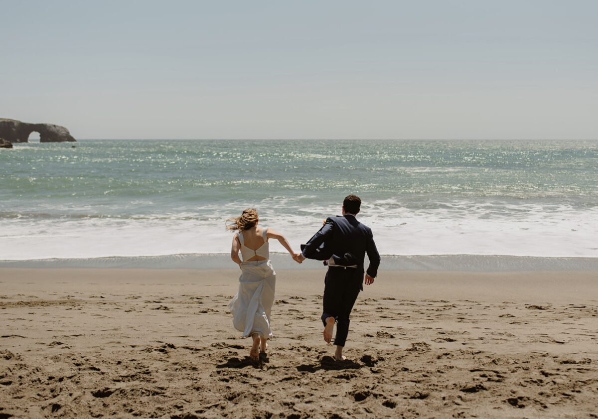 A couple in wedding clothes runs away from the camera, hand in hand toward the ocean on the beach on a sunny day. The bride wears a white dress that she holds in her free hand, and the groom wears a navy suit. Plan a multi-day California elopement with hikes, portraits, and peace. Here’s how to design an experience that feels like you.
