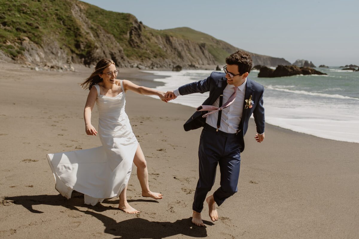 A couple runs laughing toward the camera on a sunny beach on their wedding day. The groom wears a navy suit and pale pink tie, and the bride wears a white dress with a slit in one side. In this post are gentle ways to tell family you're eloping—plus emotional tips, real scripts, and ways to honor your loved ones without losing yourself.