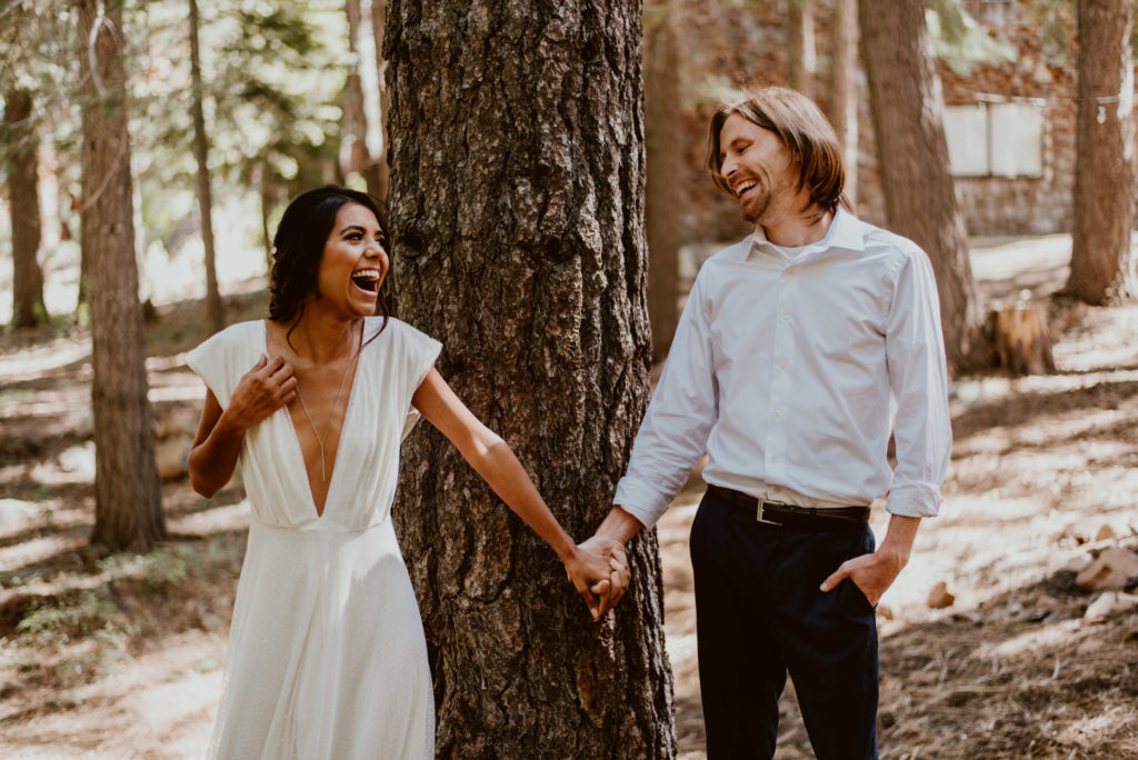 A couple looks at each other laughing while holding hands in the forest. Read on to find out how to have great photos taken of you!