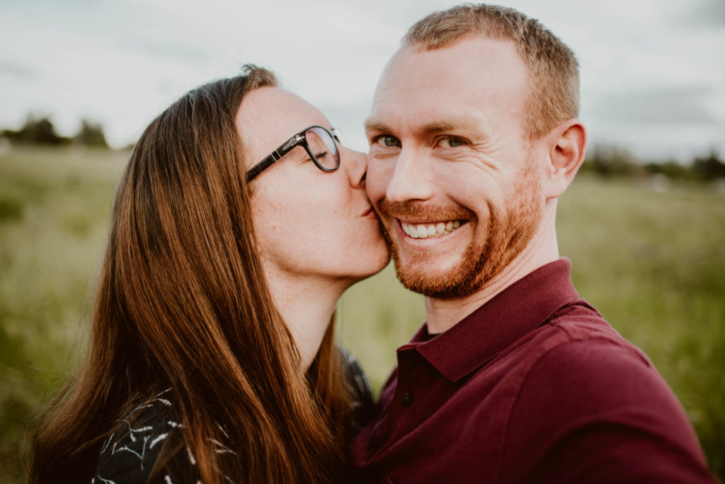A woman is kissing a man's cheek while he looks at the camera and smiles. Read this post for tips on a happy relationship!