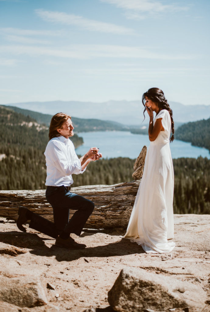 A man is down on one knee to propose. The woman is surprised and has a hand over her face. They stand on a mountain overlooking the forest and lake.