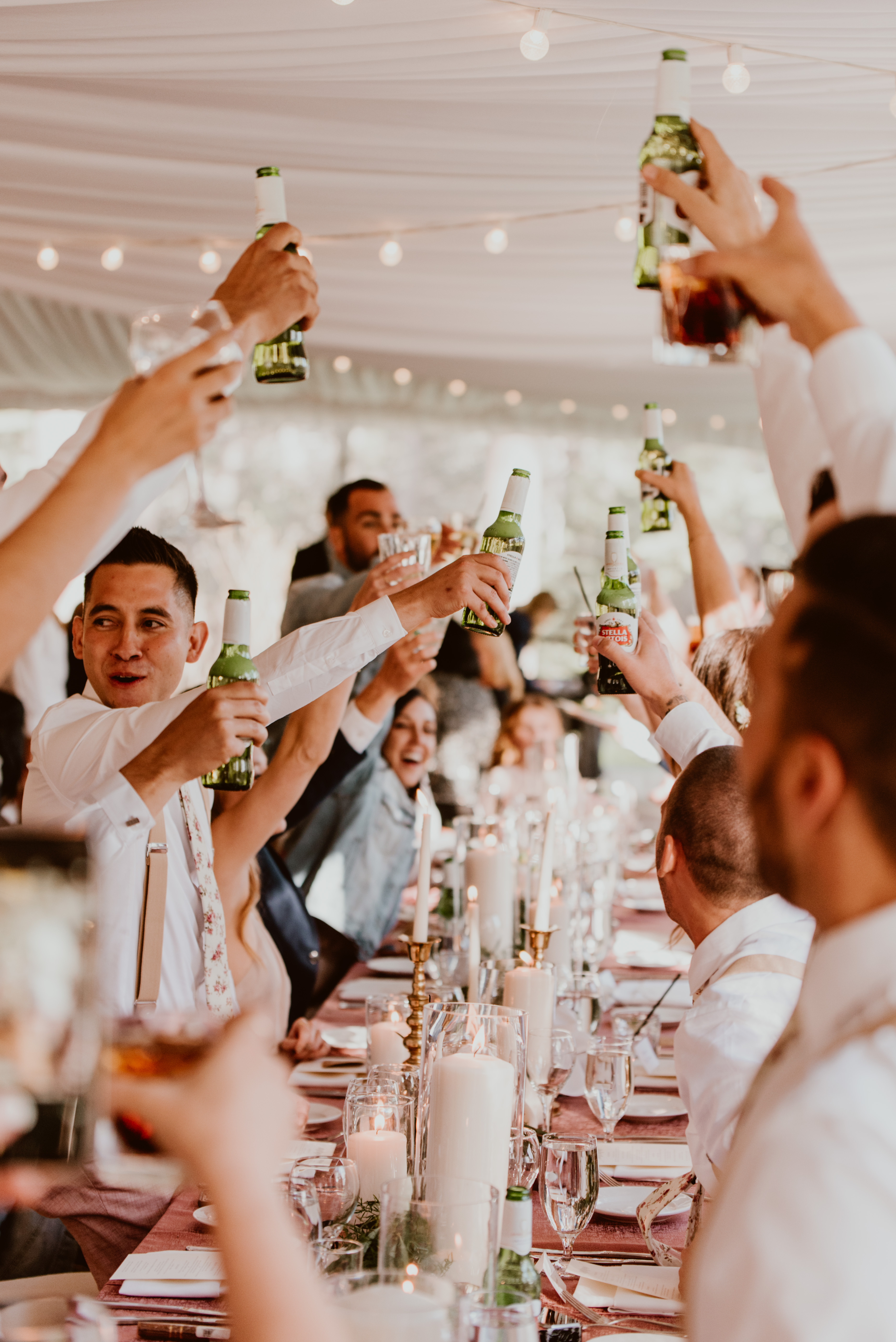 A dozen glasses are raised in a toast around a table at a wedding.