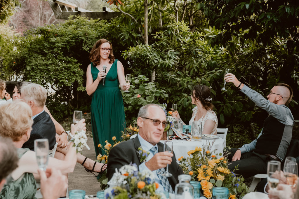 Woman is giving a speech at a wedding; the groom and bride are looking at her and glasses are raised in a toast among the guests.