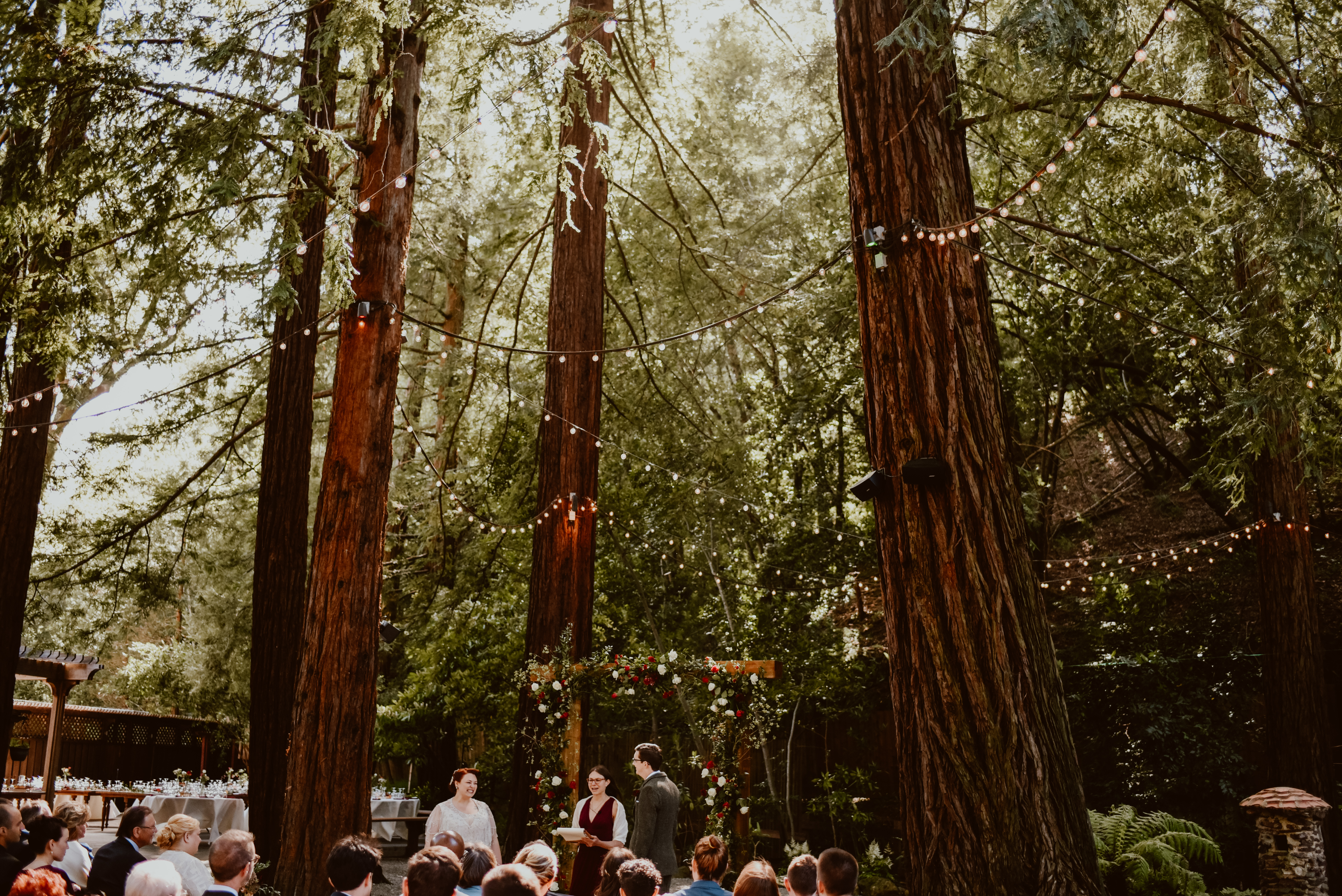 A bride and groom stand at an alter surrounded by cafe lights hung in the redwood trees
