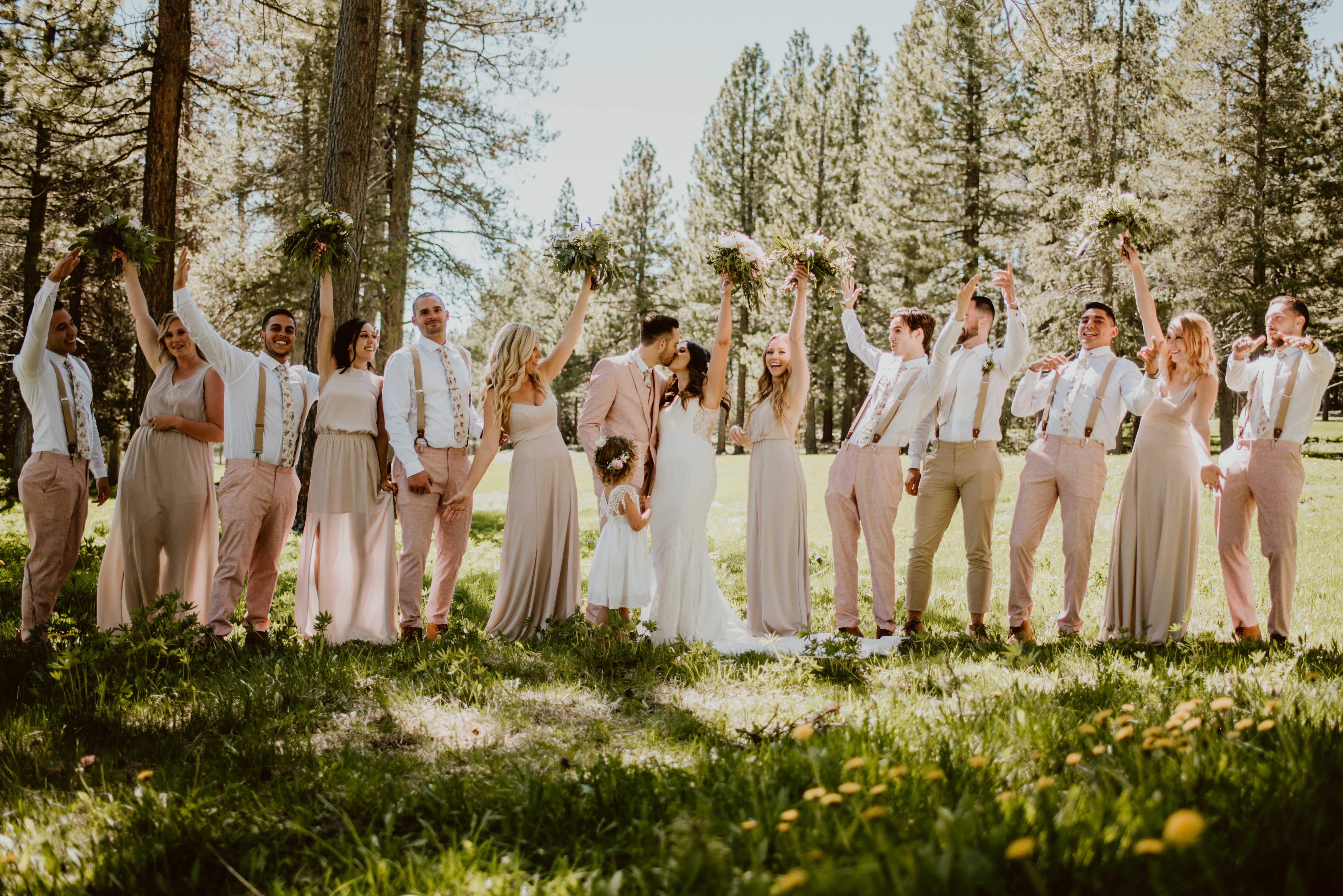 A bride and groom kiss, while their wedding party stands on both sides of them cheering. They're standing in a meadow in the forest.
