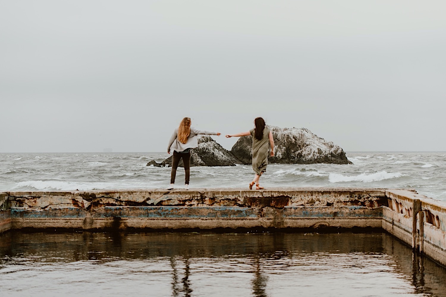 Two women with long hair stand facing away in front of the ocean with their hands outstretched reaching for each other. They stand on the ruins of the Sutro Baths in San Francisco for their engagement session. Head to this post to see more photos from my bucket list location!