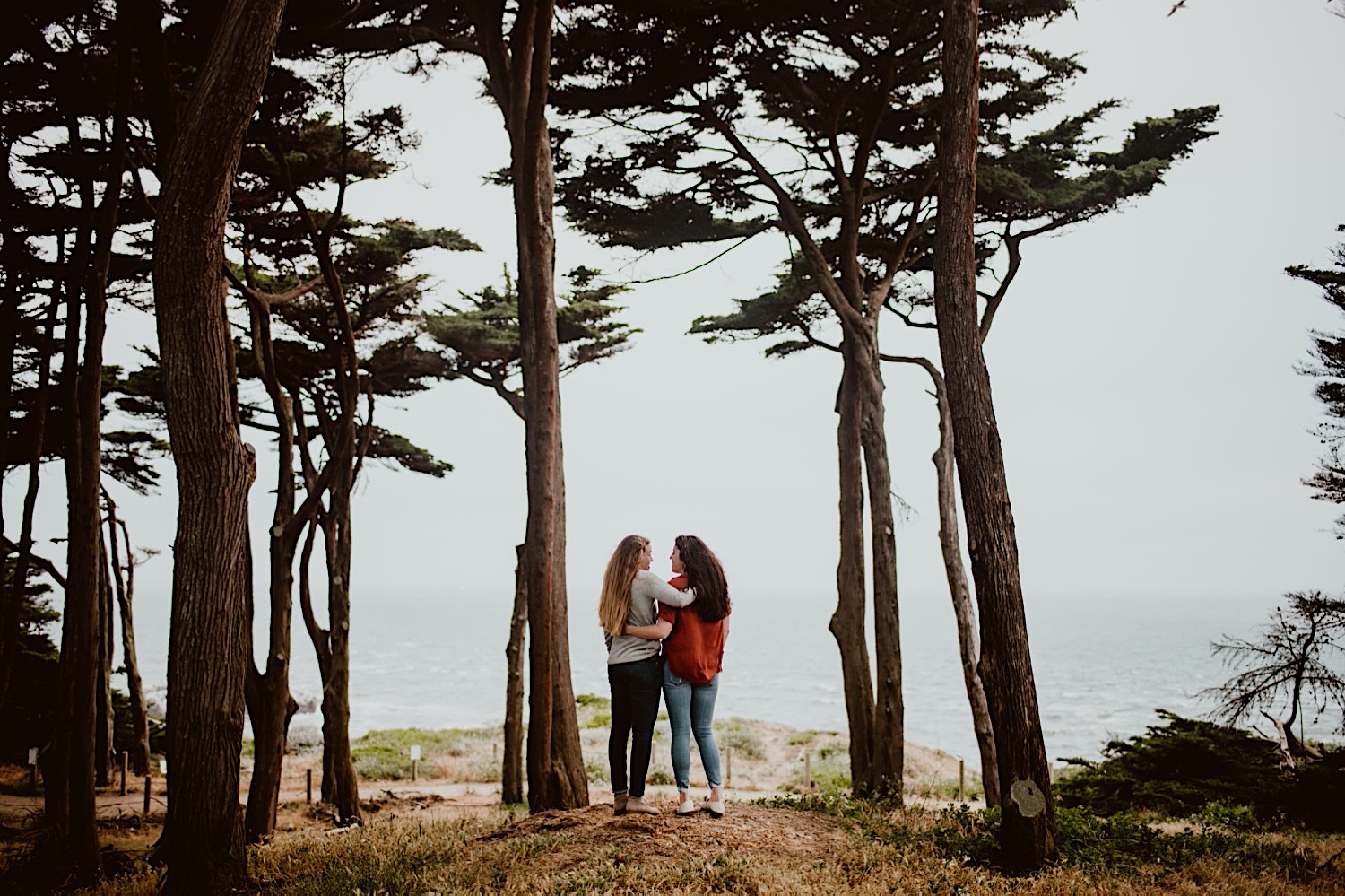 Two women with long hair are standing side by side facing away. One has an arm around the other's waist, and the other has an arm around the other's shoulder. They stand in the trees above Sutro Baths in San Francisco for their engagement session. Head to this post to see more photos from my bucket list location!