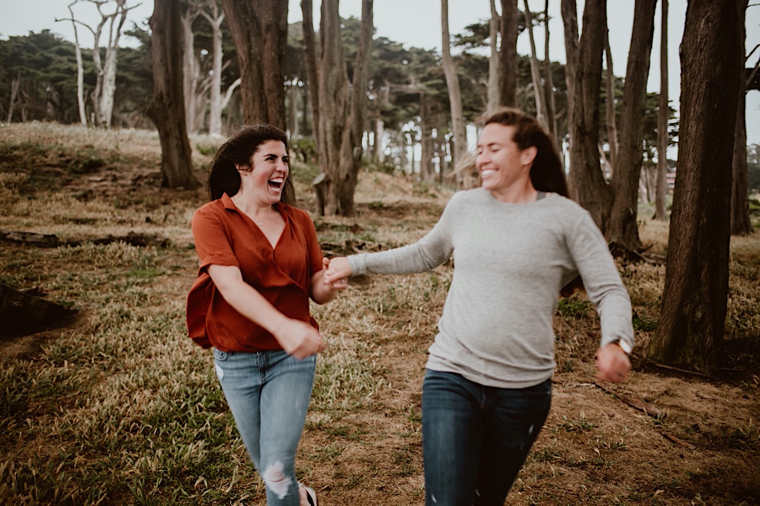 Two women are holding hands and running directly at the photographer, while looking at each other and laughing. The image is slightly blurry. They are in the trees above Sutro Baths in San Francisco for their engagement session. Head to this post to see more photos from my bucket list location!