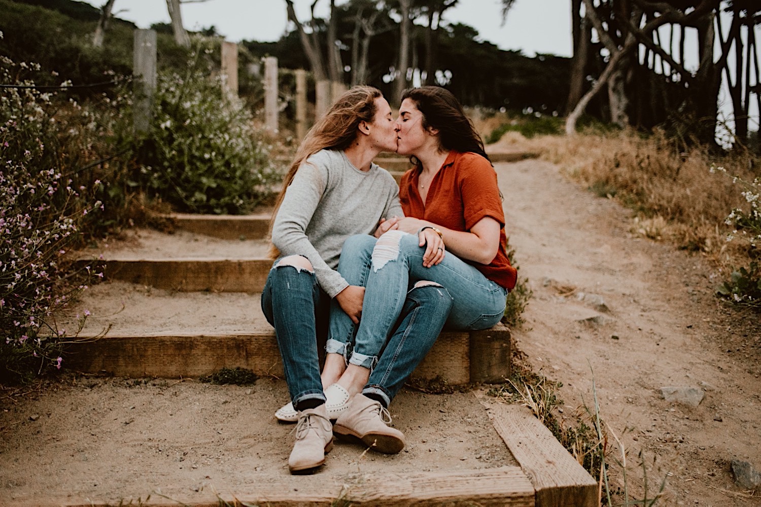 Two women sit on a set of steps built into the cliffside trail. One has her legs draped over the others' while they kiss. They sit among the trees above Sutro Baths in San Francisco for their engagement session. Head to this post to see more photos from my bucket list location!