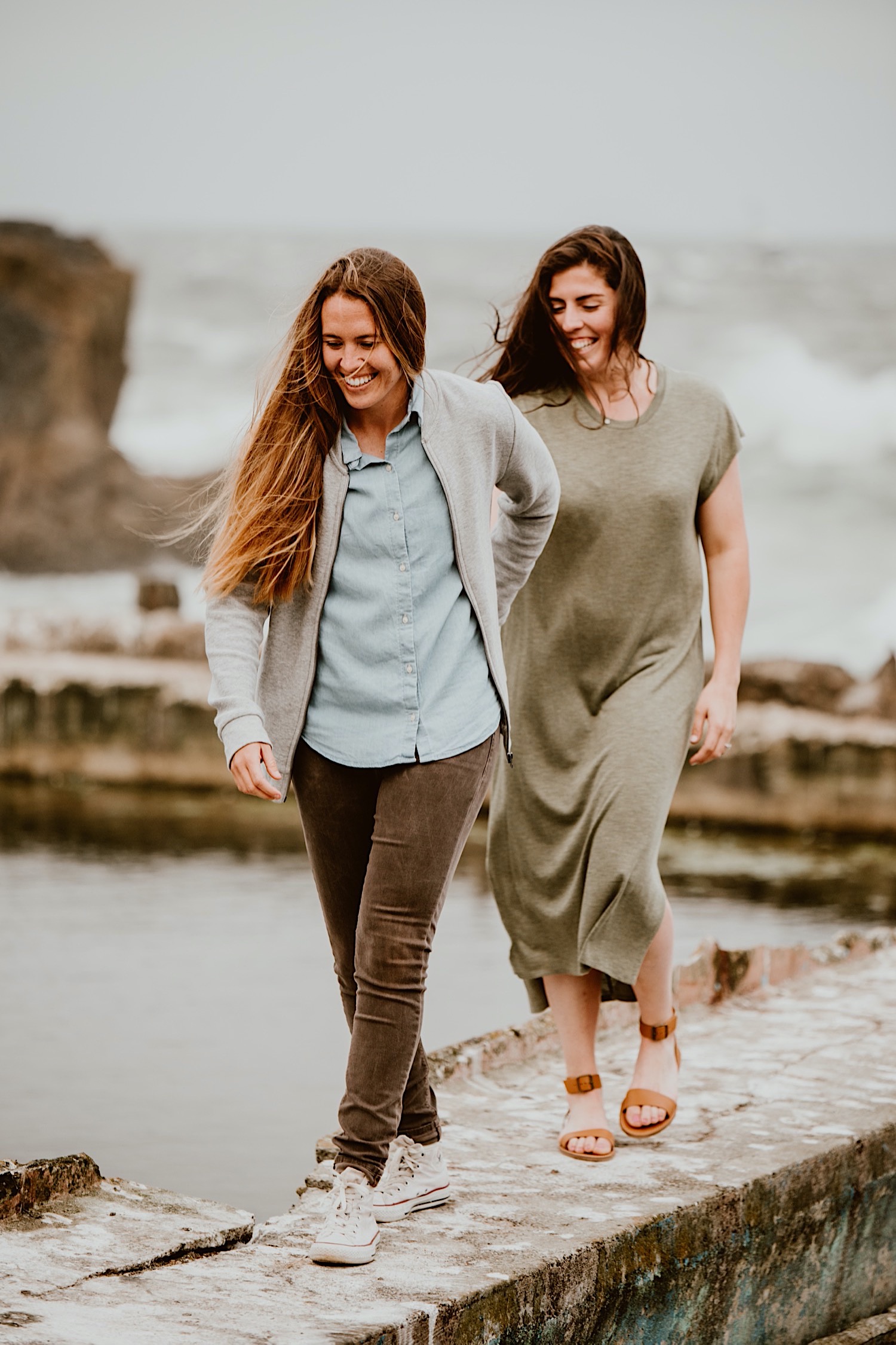 Two women with long hair are holding hands and walking single file on the ruins of the Sutro Baths in San Francisco for their engagement session; one is leading the other. Head to this post to see more photos from my bucket list location!