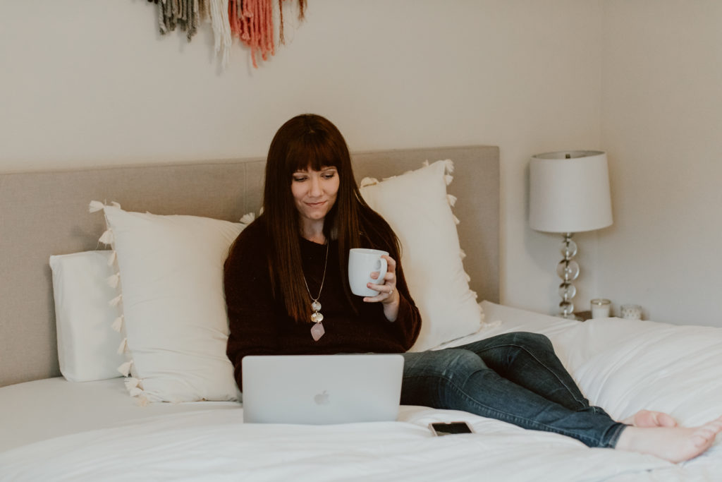 A woman lounges on a bed while working on a laptop and holding a cup of coffee. Head to this post for the top 3 tips on migrating from Honeybook to Dubsado.