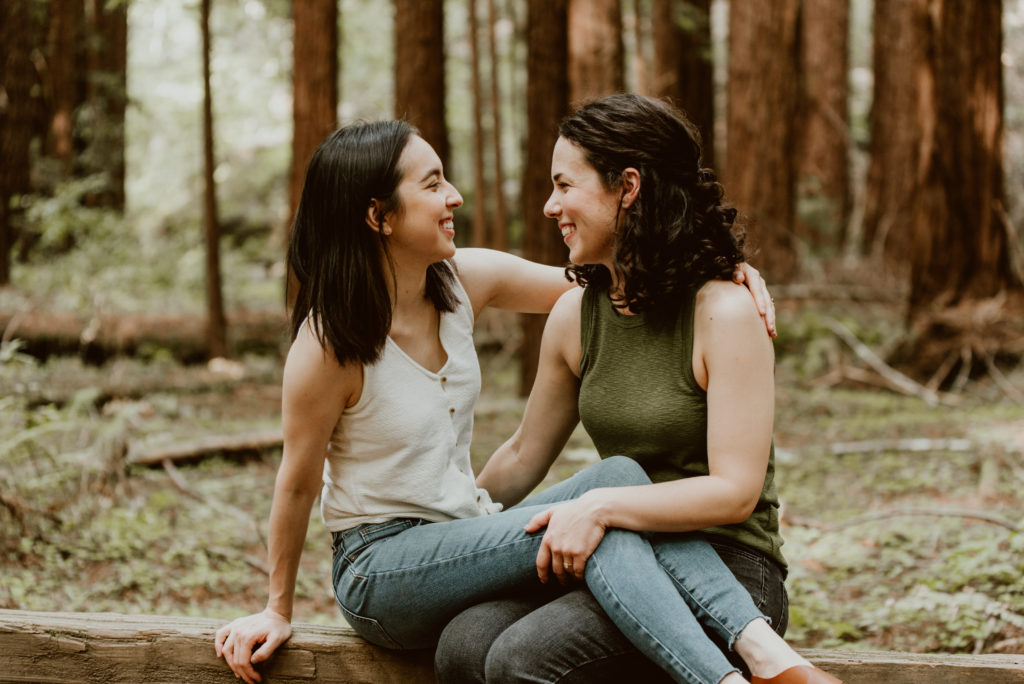 Two women sit on a bench in the forest, with one woman's legs over the other's. LGBTQ+ photos from this forest and river Guerneville engagement session