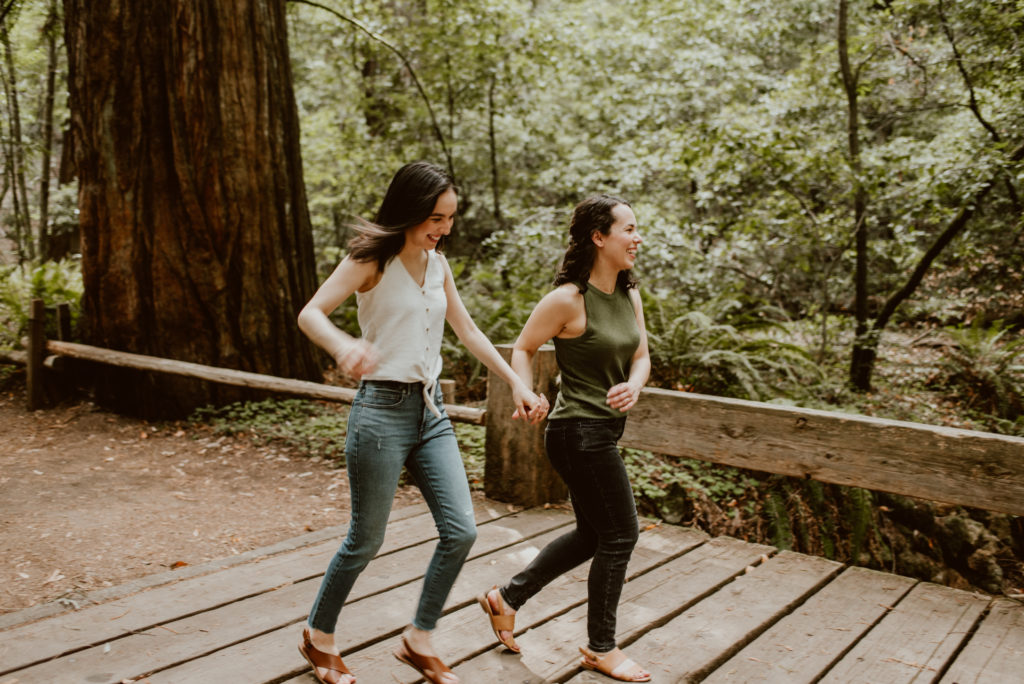 Two women run in the forest hand in hand. Be sure to check out this post for photos from their forest and river engagement session!