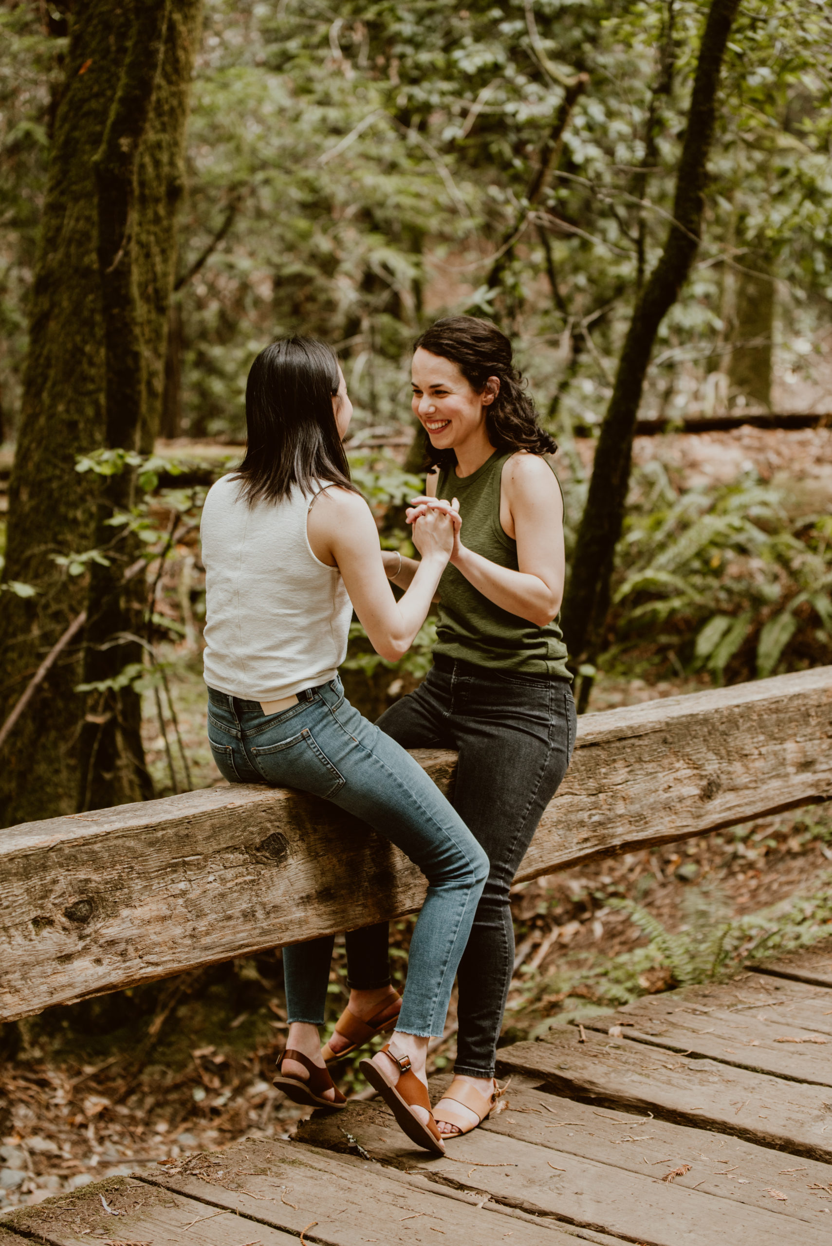 Two women sit together on a railing facing each other with their hands clasped together. Be sure to check out this post for photos from their forest and river engagement session!