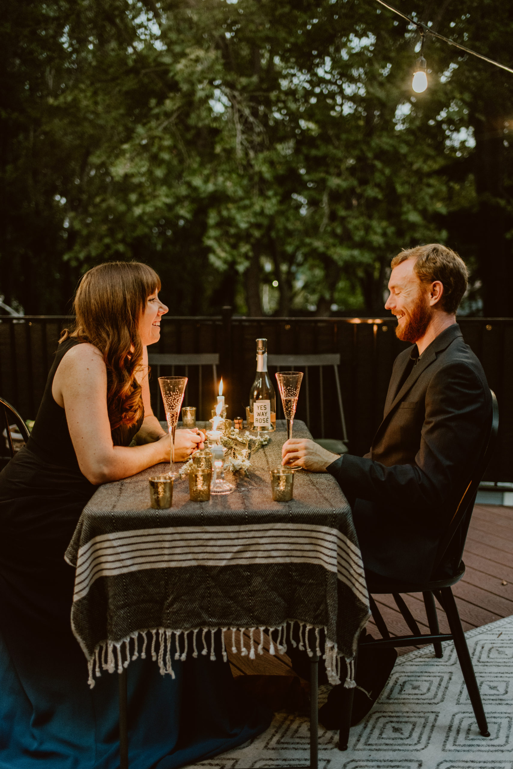A couple in formal wear sit across from each other at a table outside under a canopy of trees. Read this post for an elegant and easy way to celebrate your original wedding date.