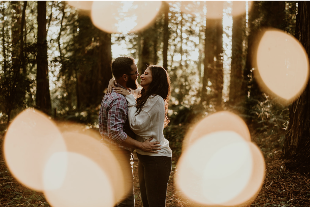 A couple embraces in the woods with a speckling of light surrounding them. If you're stuck wondering what to wear to your fall photo shoot, this guide has my five fave outfit items for your session!