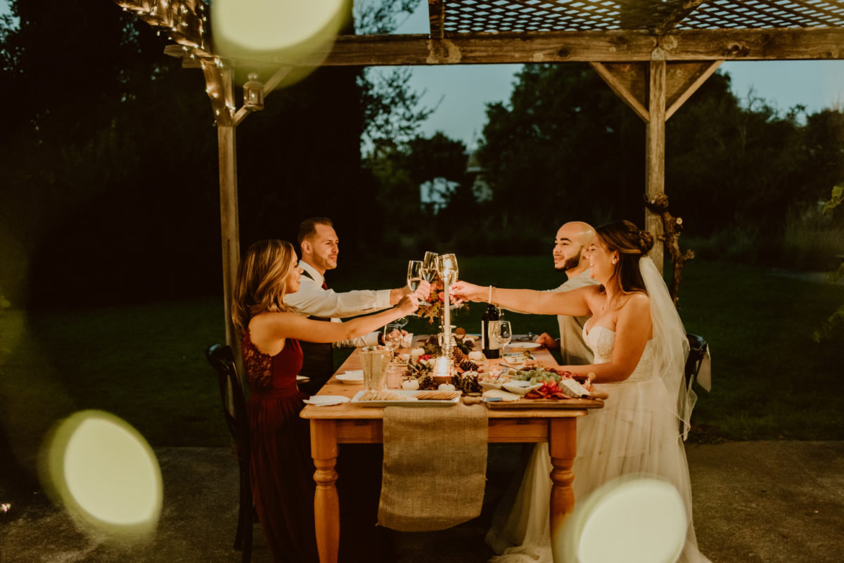 Two couples sit at a table outside sharing dinner, and are clinking glasses over candlelight. One woman is wearing a wedding dress. This page details what an elopement is and why you should have one, so be sure to give it a read if you're curious about them!