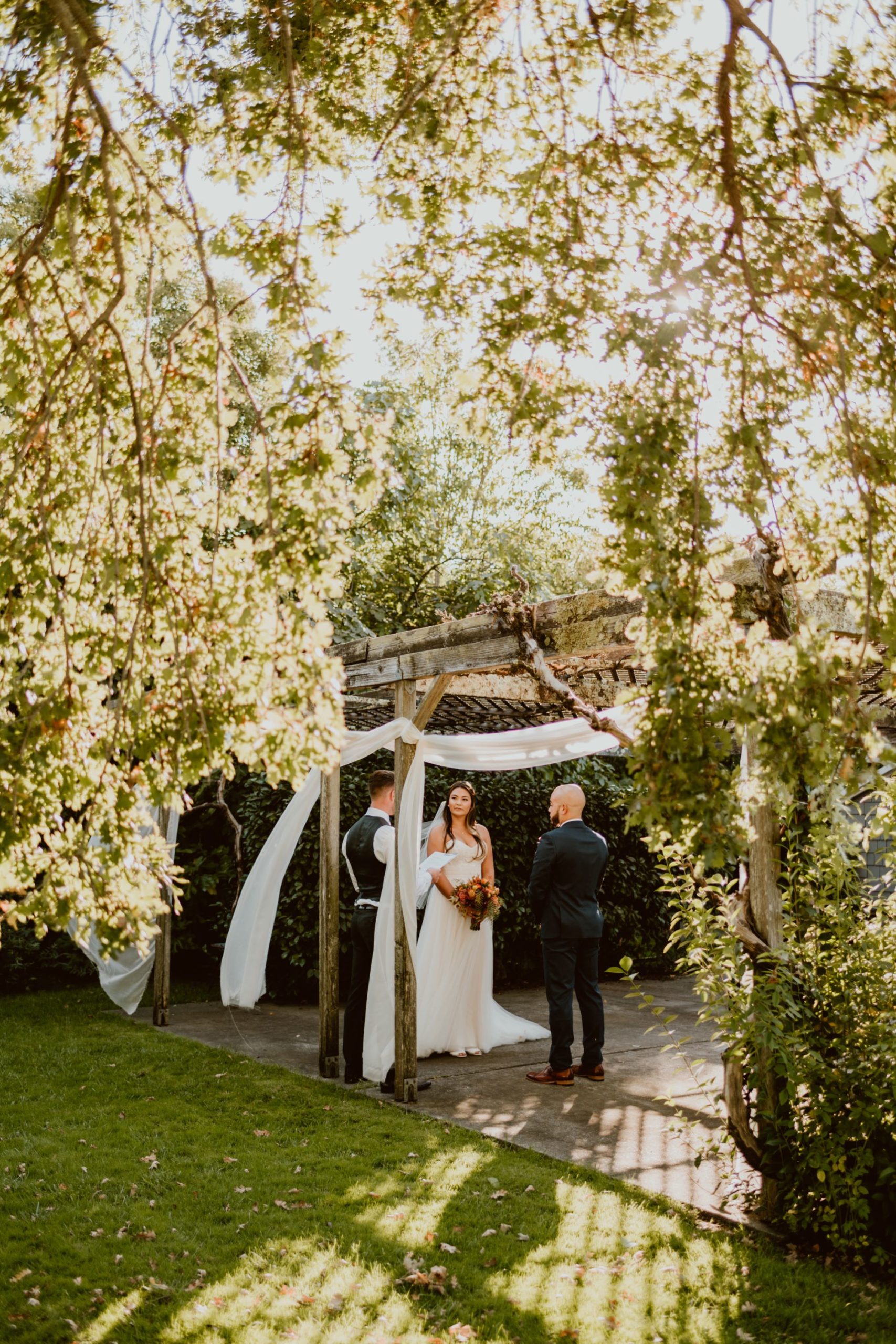 A bride and groom face each other during their elopement ceremony under a pergola and oak tree, with sunlight filtering through.

If you're currently grappling with making a Plan B or even Plan C because of covid, this backyard elopement in wine country will help give you some inspo!