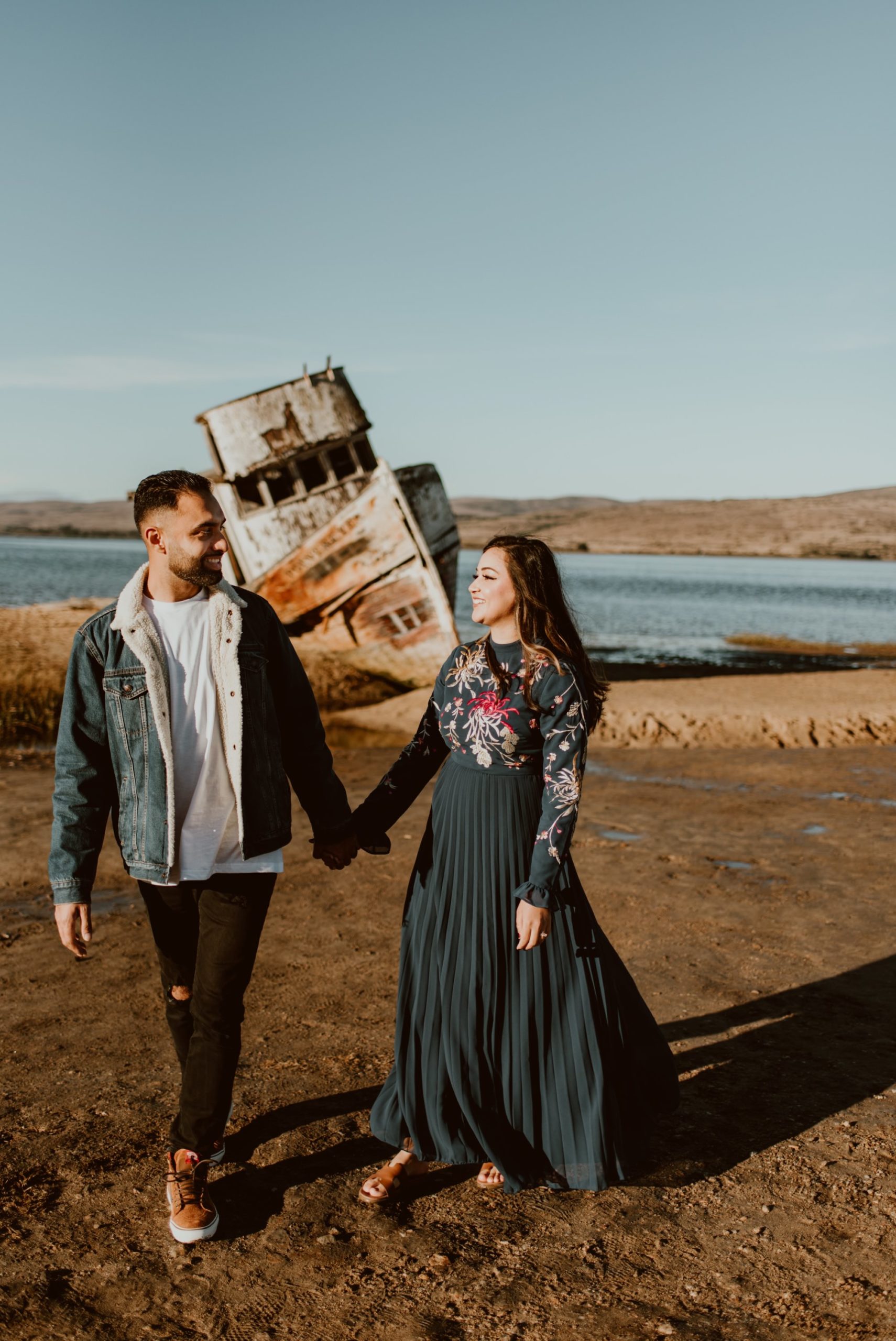 A man and woman hold hands and look at each other while they walk in front of the Inverness shipwreck during their Point Reyes engagement session.