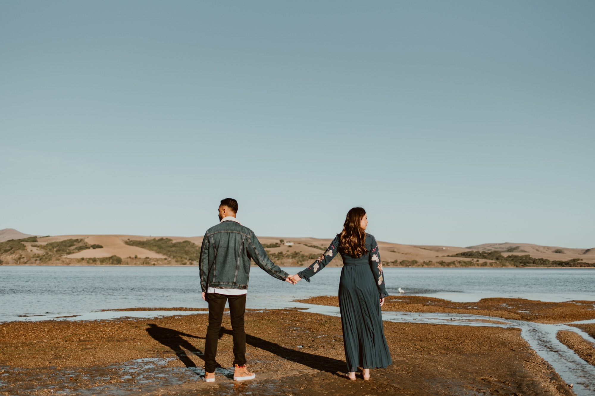 A man and woman hold outstretched hands and stand apart from each other, each looking into opposite directions in front of Tomales Bay during their Point Reyes engagement session.