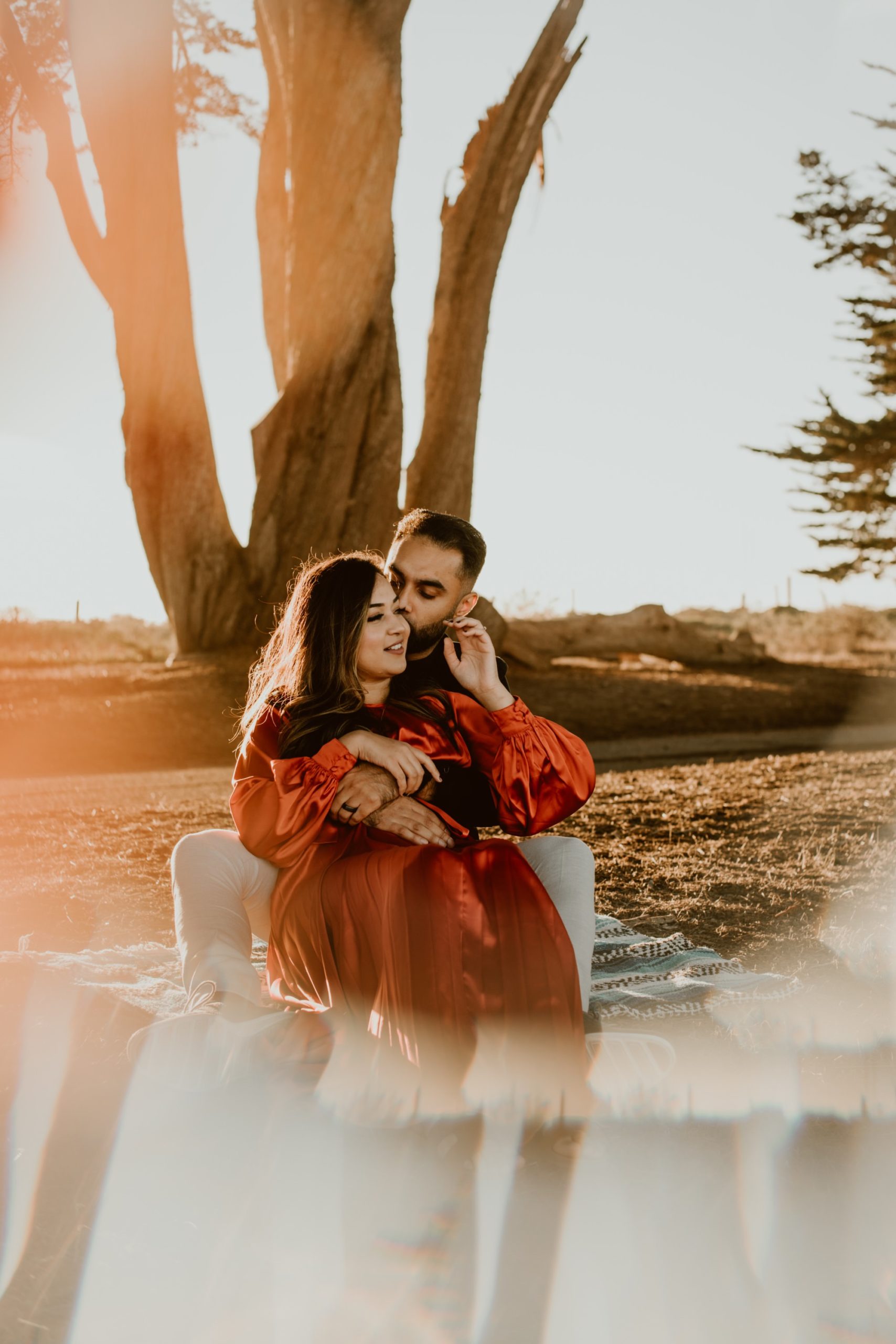 A woman sits between the legs of a man while they embrace and smile under the Cypress Tree Tunnel during their Point Reyes engagement session.