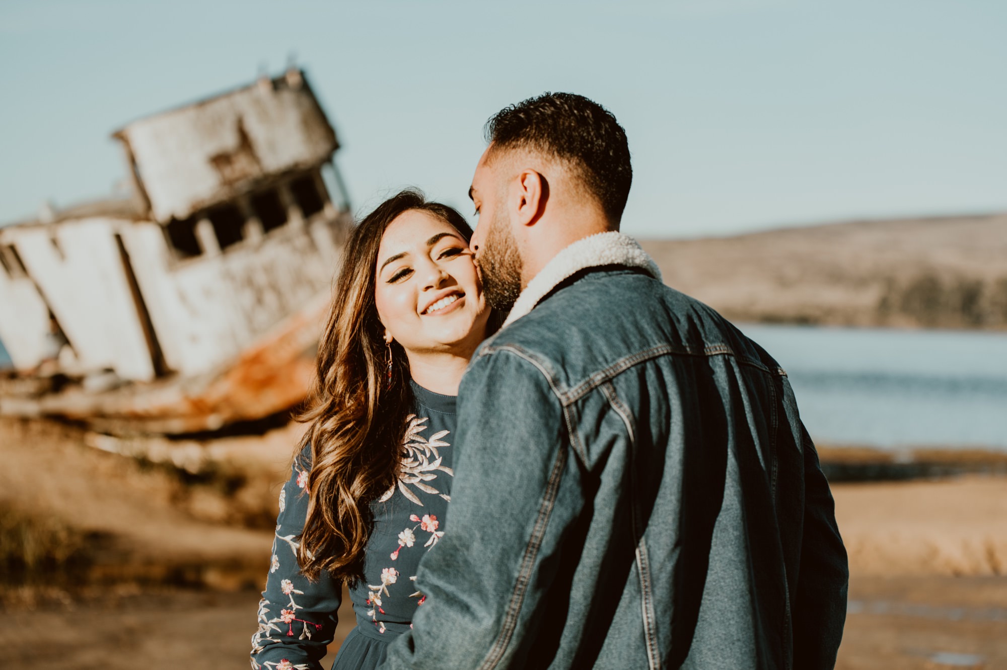 A man kisses the cheek of a woman in the sunlight while they stand in front of the Inverness shipwreck during their Point Reyes engagement session.