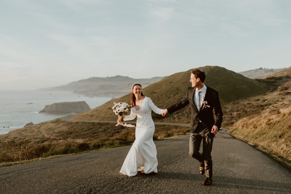 A couple run laughing hand in hand down a street on their elopement day on the Sonoma coast with the ocean in the background.