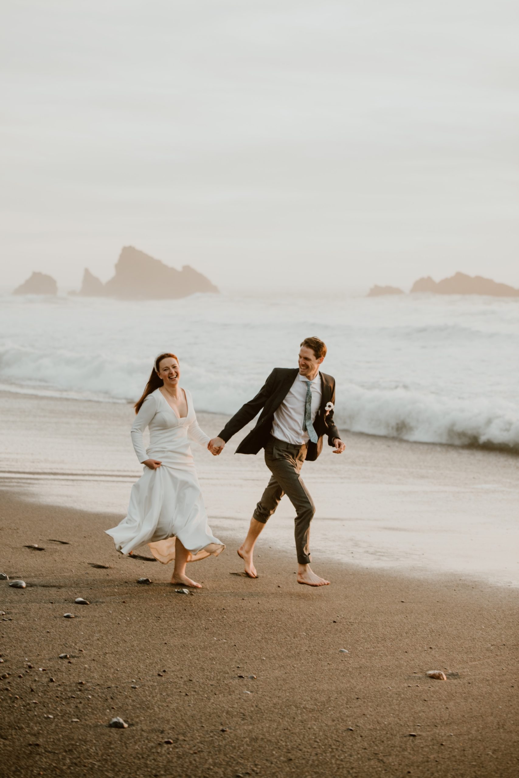A couple run laughing hand in hand along the beach on their elopement day on the Sonoma coast.
