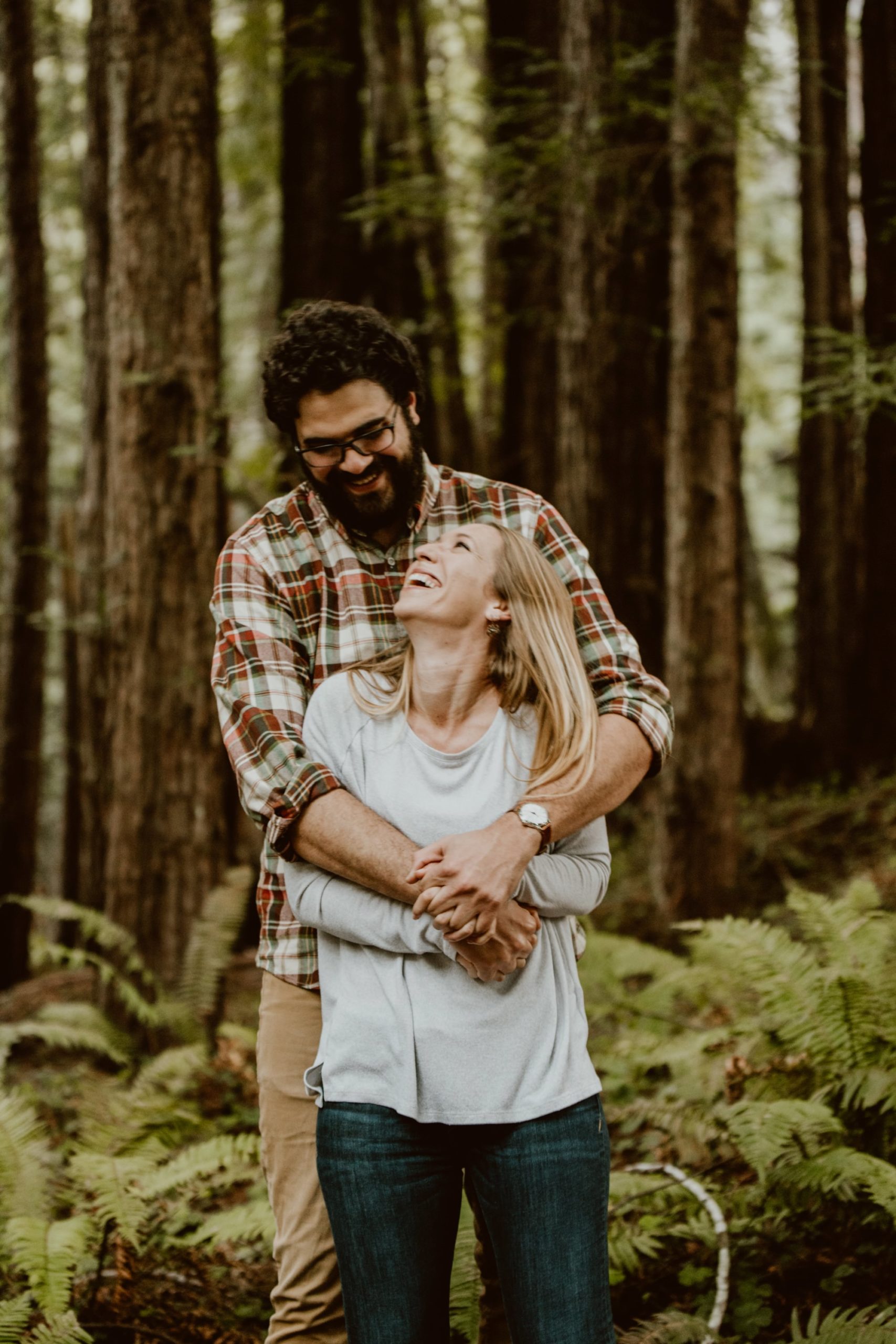 A couple stands in front of each other with arms around each other. They are looking at each other and laughing in a redwood forest on the Sonoma coast.