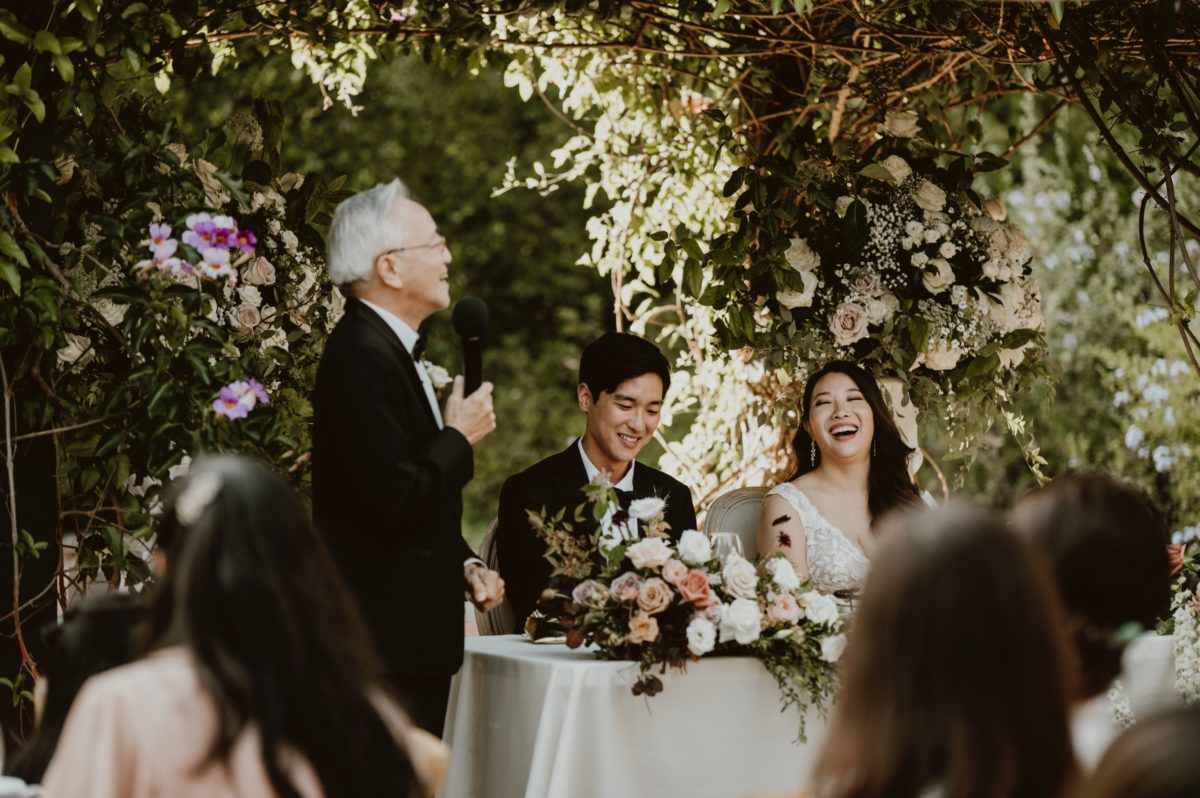 A bride and groom sitting side by side at their sweetheart table share a laugh during her dad's toast. Feeling stuck in a box with your wedding planning? Here are my 7 hot takes on wedding traditions to break you out of a planning rut.
